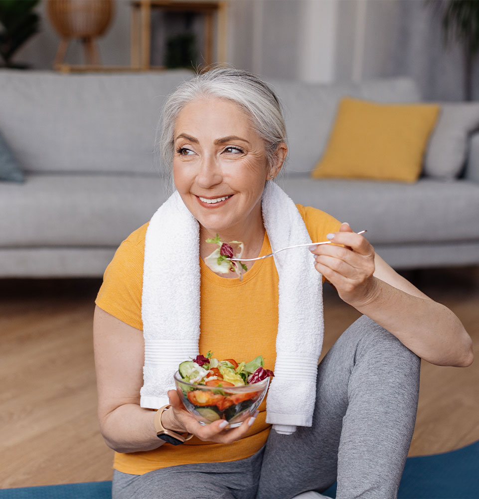 Happy senior woman eating fresh vegetable salad, sitting on yoga mat after home workout