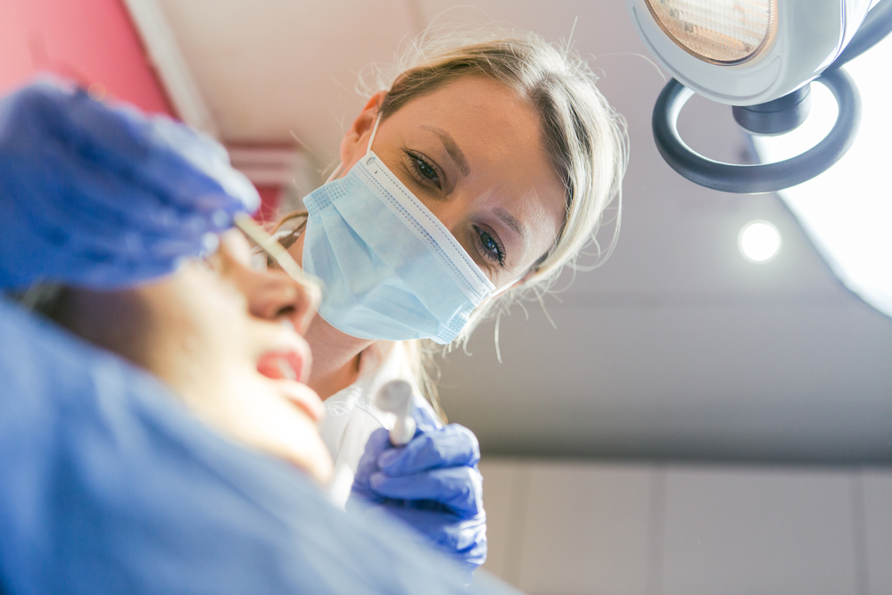 A female dentist wearing a surgical mask and gloves examines a patients mouth under a bright dental light. The patient is reclined in the dental chair, and dental tools are visible in the dentist’s hand.