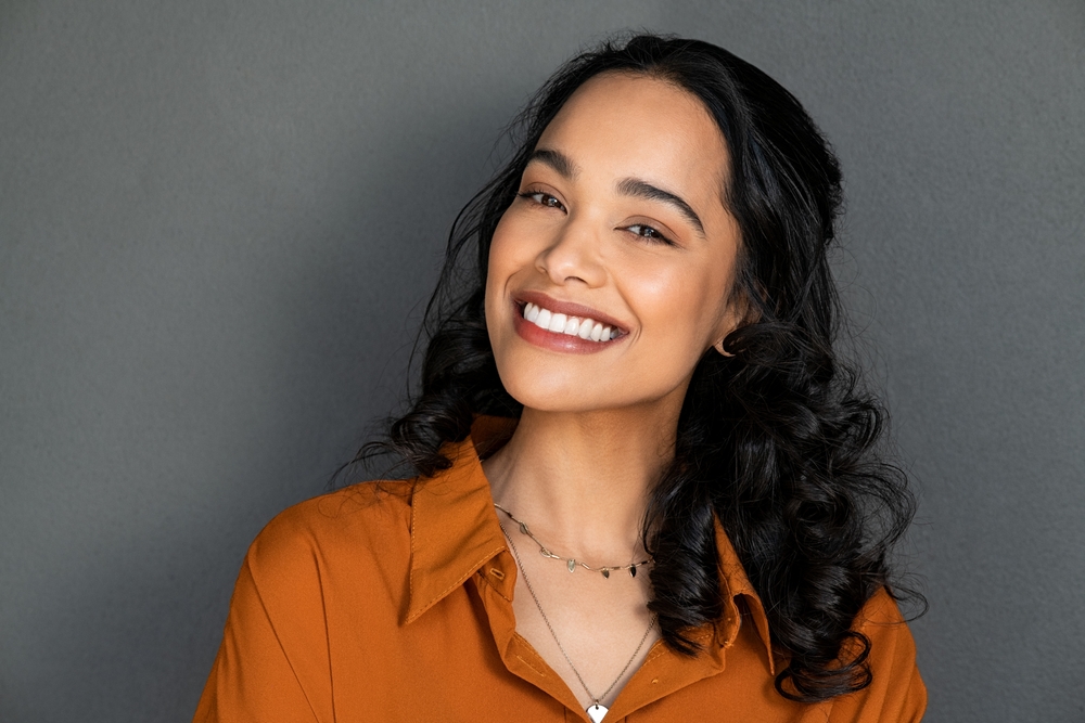 A young woman with long, wavy dark hair smiles warmly. She wears a burnt orange shirt and layered necklaces, standing against a plain gray background.
