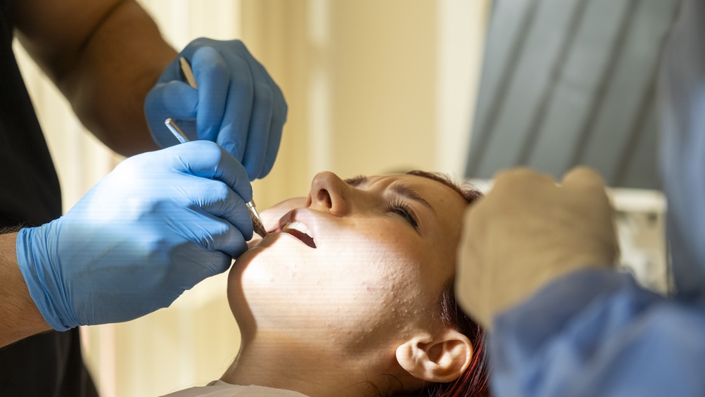 Image of a dentist placing a dental crown on a patient's tooth, with dental tools and equipment visible in the background. The patient is smiling, showing the newly crowned tooth. No text on the image.