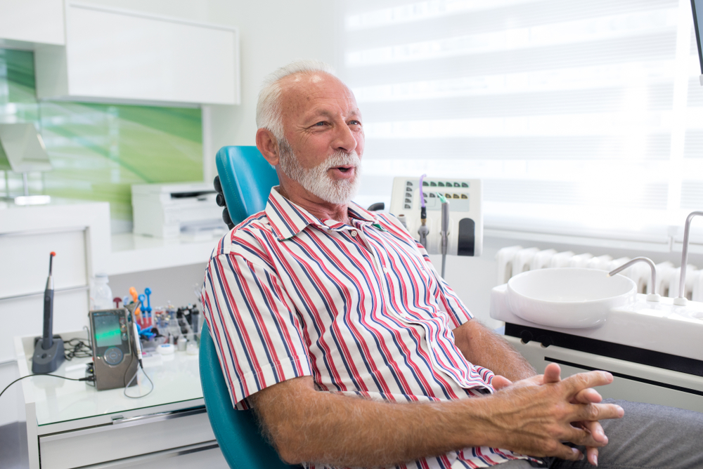 An older man with white hair and a beard, wearing a red and white striped shirt, sits smiling in a dentist’s chair in a bright office. Dental instruments and equipment are visible in the background.