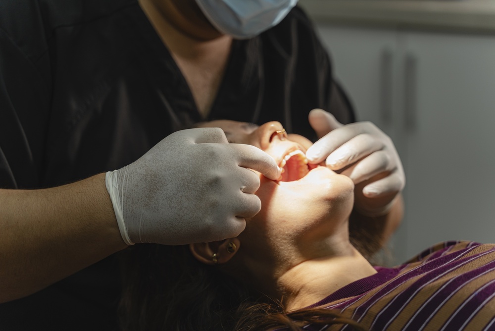 A dentist wearing gloves and a mask examines a patient’s mouth. The patient lies back in a dental chair with their mouth open while the dentist checks inside using both hands. The scene is brightly lit.